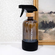Linen spray bottle with wooden cap on a white surface with a painting in the background