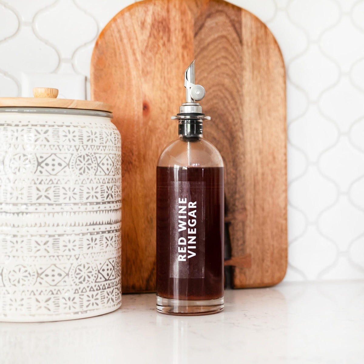 Bottle of red wine vinegar on a kitchen counter with a wooden cutting board and decorative jar.