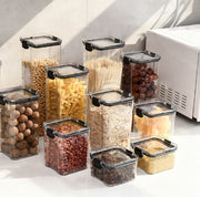 Set of clear storage containers with black lids containing various dry foods on a kitchen counter.
